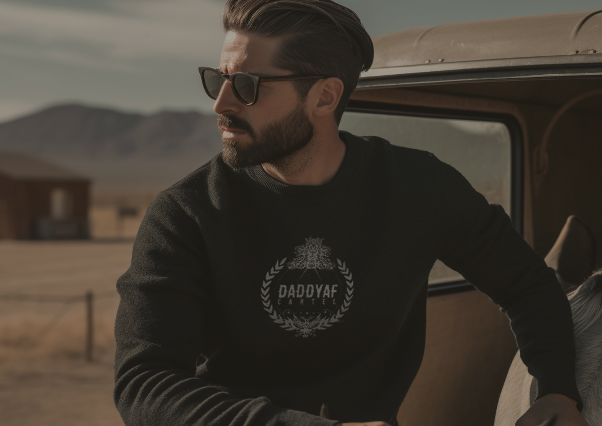 Handsome male model wearing a black cotton crew-cut DaddyAFCartel sweater with a white luxury embroidered chest Crest logo, along with classic 80’s vintage black sunglasses, outdoors on a Mexican ranch.